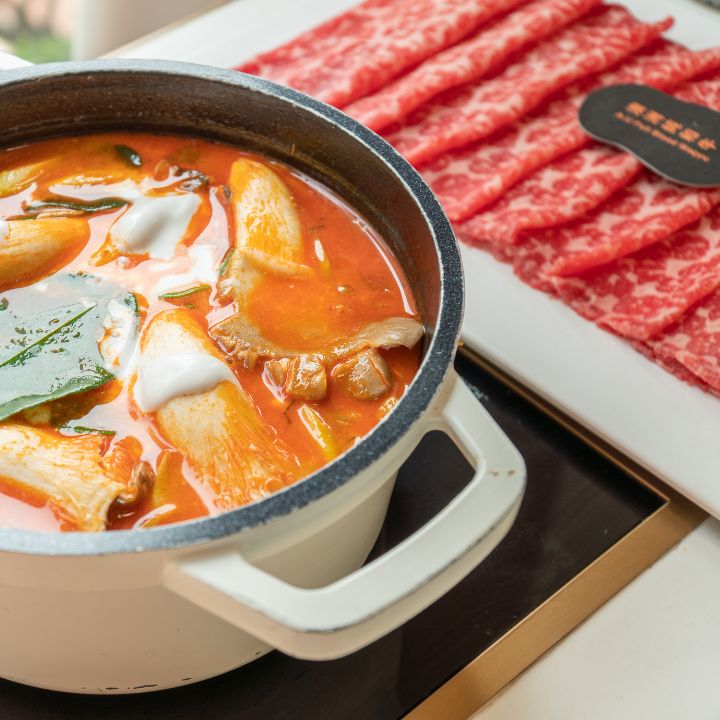 Personal spicy soup with mushrooms in white cast iron pot on single cooktop with beef slices on the side.