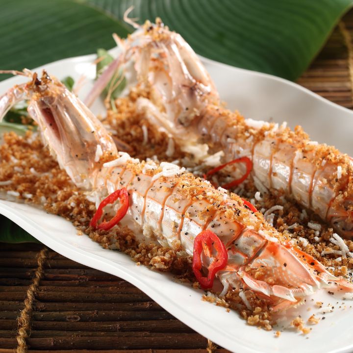 Fried light pink whole squilla with crispy garlic topping on rectangular white serving dish against thatched table cover.