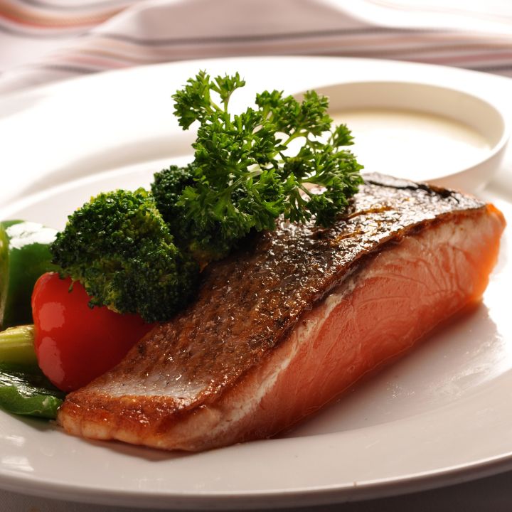 Salmon with skin on a white platter, topped with parley sprig and mixed vegetables on the side against a striped tablecloth.